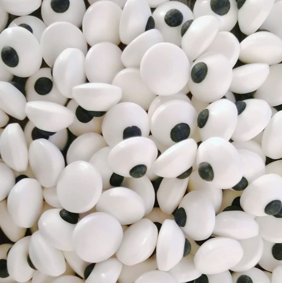 A pile of white edible candy eyes, each with a black dot in the center resembling pupils. The eyes are scattered and overlapping, creating a playful and fun texture, perfect for topping ghost-themed Halloween sandwiches.